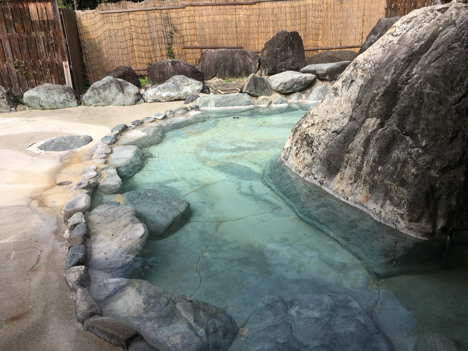 白馬八方温泉おびなたの湯の露天風呂。透明な青緑の湯と、湯が流れた跡が白く積もった巨大な一枚岩、葦簀と板塀