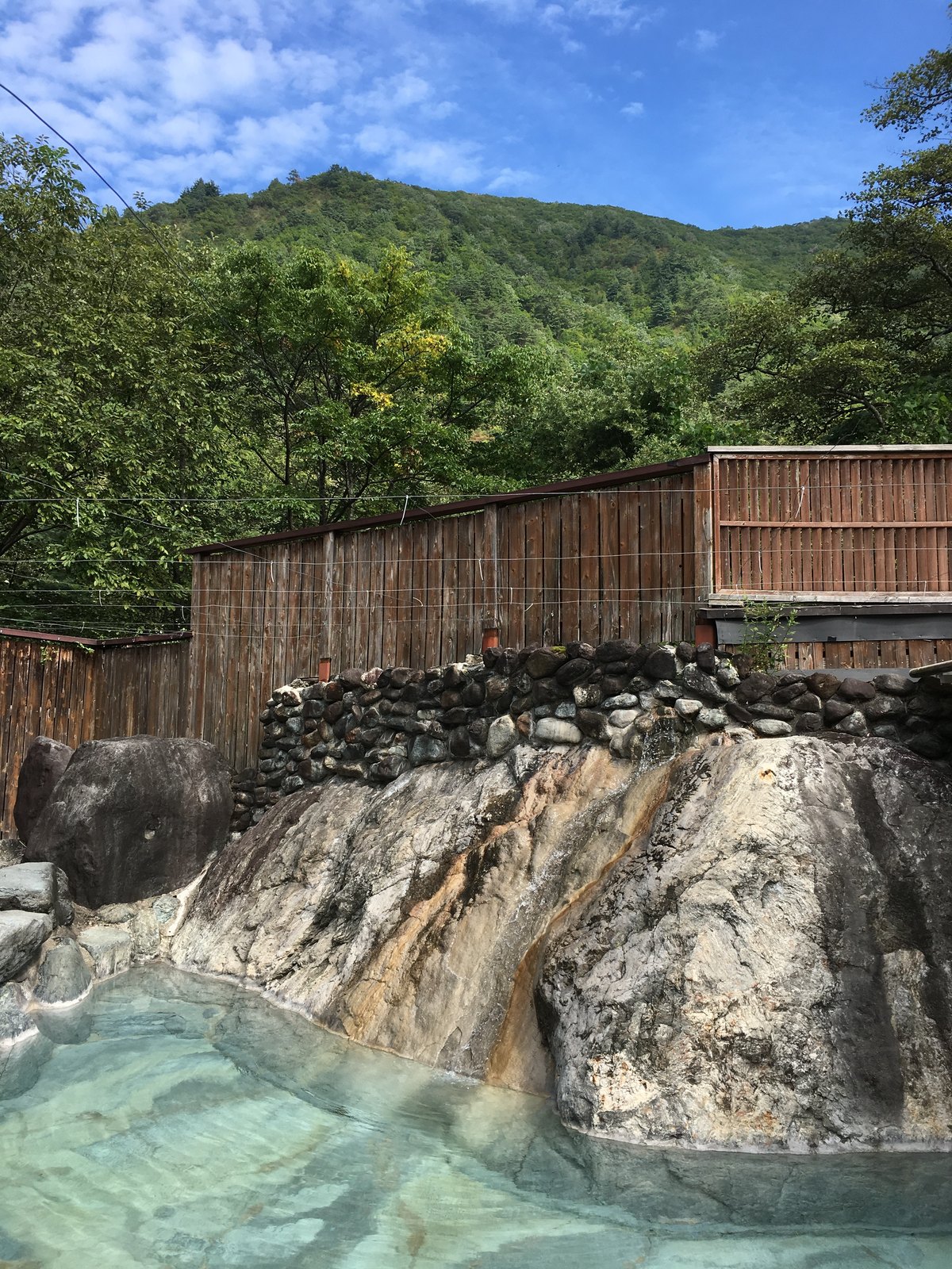 おびなたの湯の露天風呂。湯船のすぐ横にせり出した大きな一枚岩と、その上の石積みと板塀、背後に緑の山と青空